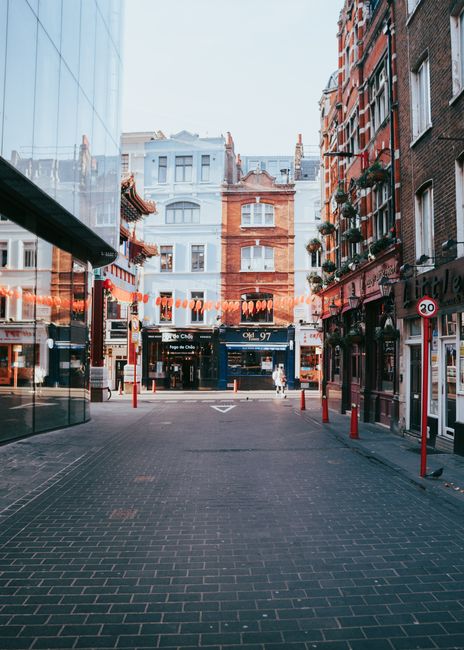 A quiet urban street scene in Soho featuring a paved pedestrian area with dark grey bricks. On the left side, a modern glass building with reflective windows extends outward, partially framing the scene. On the right, several multi-storey brick buildings with traditional façades, some decorated with flower boxes and hanging plants, line the street. In the background, a large, ornate, white and terracotta-colored building dominates the view, with additional older buildings visible behind it. String lights hang across the street mid-height, adding a decorative element. The street is bordered by red bollards and signs indicating a maximum speed limit of 20 miles per hour, subtly connecting to local traffic and waste collection regulations. The scene is evenly lit, with clear skies overhead, creating a calm atmosphere typical of a city centre environment that occasional private waste collection services, like those offered by Rubbish Removal Soho, might operate within.