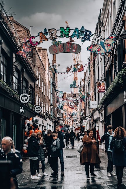 A bustling pedestrian street during daytime, decorated with colorful carnival-themed banners and ornaments hanging overhead, including butterflies, balloons, and streamers. The street is lined with storefronts on both sides, featuring modern signs and window displays, with people casually walking and engaging in conversation. Some visitors are holding umbrellas, indicating overcast weather. In the foreground, a few individuals are standing and interacting, while others are walking further into the scene. The ground appears wet, reflecting the urban environment and decorations. Behind the storefronts, brick and painted facades are visible, and above, the sky is filled with dark clouds, suggesting impending rain. The overall scene captures a lively, decorative urban setting typical of a busy shopping or entertainment district, ideal for local waste management and rubbish removal services that operate in such active areas, including those provided by Rubbish Removal Soho.