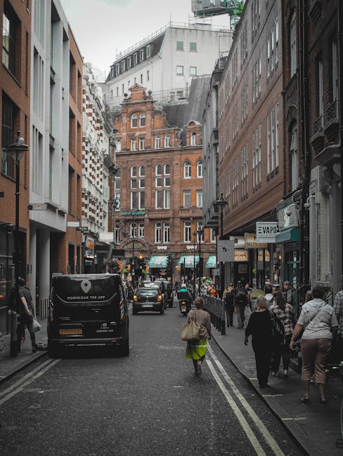A narrow city street in an urban environment, flanked by tall multi-storey buildings on both sides. The buildings feature a mix of modern and historic architectural styles, with some displaying ornate brickwork and decorative windows, and others with sleek glass facades. The street is paved with dark asphalt, and double yellow lines run along the edges. On the left, a black van with company branding is parked, and on the right, several pedestrians are walking along the sidewalk, some carrying bags or talking. Shopfronts with signage and awnings are visible on both sides, including a shop called 'E vapor' on the right. In the background, there are more commercial buildings, with a larger, more ornate brick building visible at the end of the street. The lighting suggests an overcast day with diffuse natural light, and the scene captures typical urban activity related to shopping or commuting, illustrating a setting where various waste collection and rubbish removal services could be relevant in this busy city environment.