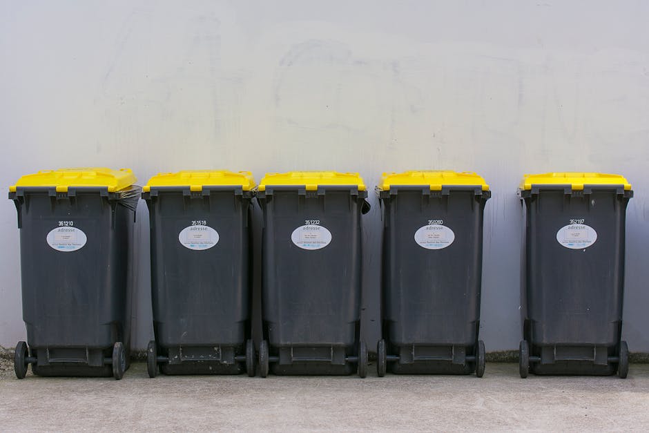 A row of five large wheeled rubbish bins made of dark grey plastic with textured surfaces, positioned against a plain, light-colored wall. Each bin has a bright yellow lid with a ridged finish, and white oval labels with identification numbers affixed to the front. The bins are situated on a concrete pavement, aligned neatly in a straight line, with their wheels visible at the base. The environment appears to be an outdoor or semi-outdoor space, possibly within a residential or commercial service area. The scene reflects a typical setup for waste collection or private rubbish removal, with the bins ready for collection by a waste management service such as Rubbish Removal Soho, offering an alternative to council-led rubbish disposal services. The lighting is neutral, highlighting the clean and organized appearance of the rubbish storage system, emphasizing the importance of proper waste handling for both residents and local businesses.