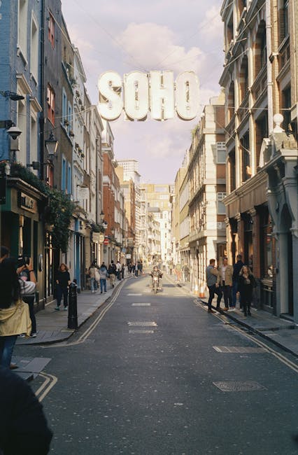 A view down Wardour Street in Soho, W1F, featuring a narrow pedestrianized roadway lined with multi-story commercial buildings on both sides. The buildings display various materials, including brick, stone, and painted facades, with some featuring decorative architectural details such as cornices, window trims, and balconies. The street is populated with pedestrians walking along the sidewalks and crossing the road, some engaged in conversations or using mobile devices. In the background, large white letters spell out 'SOHO' suspended above the street, hanging from wires attached to the buildings. The sky overhead is partly cloudy, with soft natural light illuminating the scene, creating shadows on the pavement and building facades. This setting reflects an urban environment suitable for public and private waste collection, typical of the area where independent rubbish removal services may operate to manage waste generated by shops and restaurants, aligning with non-local-authority disposal options generally offered by companies like Rubbish Removal Soho.