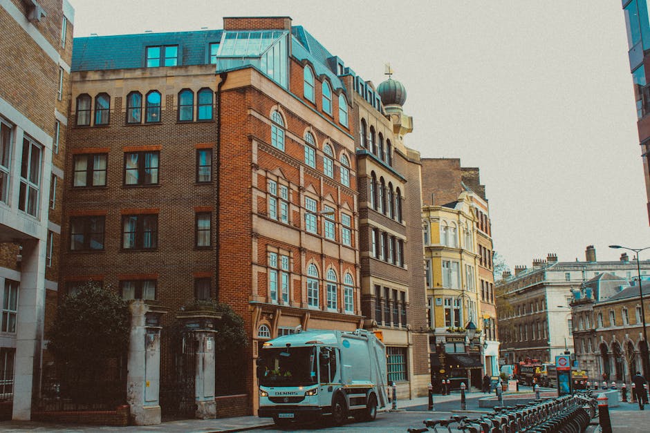 A street scene in an urban area showing a variety of historic and modern buildings lining the road. In the foreground, a light blue waste collection vehicle is parked on the pavement next to a row of black metal bike racks. The vehicle has a rounded cab and a compact body, typical of recycling or rubbish collection trucks used for private waste removal services. Surrounding the vehicle, there are small trees near an entrance gate made of stone columns and metal fencing, indicating a gated property or estate. The buildings feature diverse architectural styles with brick facades, large arched windows, and a turret topped with a decorative onion dome. The scene is set during daylight with overcast weather, providing even natural light that highlights the textures of brickwork, metal, and glass. The street is relatively quiet, with a few cars and pedestrians visible in the distance, reflecting a typical city environment suitable for private rubbish removal and on-site clearance activities by companies like Rubbish Removal Soho.